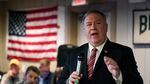Mike Pompeo holds a microphone in his right hand while he speaks to a room of people with an American flag on the wall in the background. Maskless people are visible in the background.