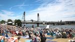 People sit on picnic blankets at a waterfront park in Portland, while boats gather on the river and a bridge is visible in the background.