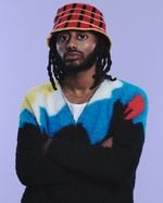 A man in a bucket hat and sweater stands with arms folded.