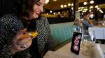 A woman sitting at a restaurant holds a drink and has a video call with a man shown on the screen of her smart phone that is propped up by a glass.