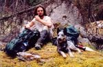 Ein Mann sitzt auf einem bemoosten Waldboden mit einem Rucksack in der Nähe und einem Hund zu seiner Linken.