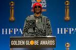 Snoop Dogg wears a red hat and a black and white patterned shirt as he stands at a podium that has the words "Golden Globe Awards written
