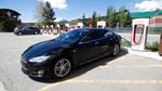 A black Model S Tesla sedan charges at a Tesla supercharging station on a sunny day.