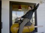 A person in a black beanie, black sweatshirt, and gray pants rides a mechanical corn dog.