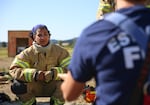 A firefighter crouches and faces the camera while another person's back is visible.