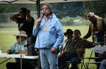 A man in braids wearing a blue collared shirt that has small feathers printed on it speaks into a microphone. Other tribal members sit behind him.