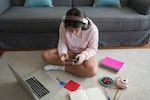 A girl sits on a rug while viewing a smartphone and wearing headphones. An open laptop is on the rug in front of her, and notebooks and pens are also on the rug.