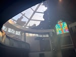 The roof of the sanctuary in a church is completely burned through, showing the exposed sky. A colorful piece of stained glass feature a cross remains in tact.