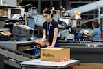 In this photo, a young woman wearing a black T-shirt attaches a label to a cardboard box. Near her is a conveyor belt.