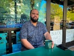 A Black man sits on a patio table with a green mug of coffee. A glass storefront is behind him, on which there are signs that read: "BLACK LIVES MATTER."