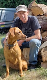 A man sits in front of a pile of cut logs with an orange-ish brown dog next to him