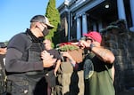 A man wearing a mask and all-black padding clothing that appears to be a bulletproof vest holds his hands near his chest, as another man gestures excitedly at him while not wearing a mask.