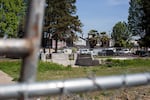 This photo shows an empty lot covered by grass of varying thickness. One part of the lot has two partial cinder block walls. In the background is a row of two-story homes.