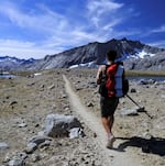 A hiker carrying a backpack looks towards a dusty trail, mountains and blue sky.