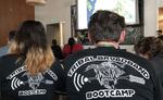 Two people sit in a room with their backs to the camera. The backs of their black T-shirts read "Tribal Broadband Bootcamp" in white letters.