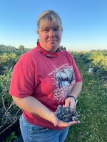 A woman stands outside in a field and holds a cluster of blueberries in the palm of her right hand.