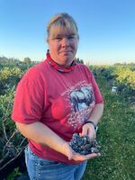 A woman stands outside in a field and holds a cluster of blueberries in the palm of her right hand.