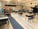 An adult wearing a mask stands in an empty classroom where tape on the floor marks off distances between student desks.