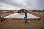 Anna Perks, wearing blue jeans and a brown jacket, walks past a large rectangular slab on the ground — the last remaining part of an old farmhouse. The slab is surrounded by flat, brown dirt ground that's punctuated by large piles of debris.