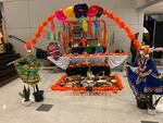 An altar with photos and candles sits in a high school common area. In the foreground is a flowered arch with skeletons in traditional Mexican dresses on each end.