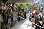 A line of National Guard troops in helmets and with shields stands on steps of a federal building with a large crowd of protestors a few feet in front of them, one protester holds a large American flag. This took place on Saturday in Los Angeles during a "No Kings" protest.