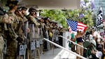A line of National Guard troops in helmets and with shields stands on steps of a federal building with a large crowd of protestors a few feet in front of them, one protester holds a large American flag. This took place on Saturday in Los Angeles during a "No Kings" protest.