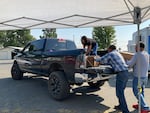 A woman stands on the bed of a large pick-up truck moving boxes as two men help her load boxes onto the truck.