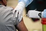The gloved hands of medical practitioner, who is standing just off camera, injects the vaccine into a man's upper right arm.