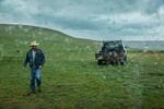 Nash walks through a verdant green field in front of an old pickup truck. Rain drops splotch the lens as gray storm clouds roll through the sky.