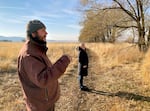 A man and woman stand on a nature trail backgrounded by bunchgrass and mountains.