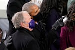 Barack Obama, on the right, wraps his right arm around George Bush in an embrace while he points to something they're both looking at. Both are wearing face masks and coats.