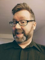 A man in a black button-up shirt and glasses smiles at the camera.