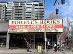 An exterior shot of the entrance to Powell's City of Books on a sunny day. The store's marquee advertises several upcoming author events, and an employee is down below with a pole used to switch out the letters.