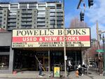 An exterior shot of the entrance to Powell's City of Books on a sunny day. The store's marquee advertises several upcoming author events, and an employee is down below with a pole used to switch out the letters.