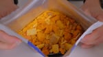 A pouch for a freeze-dried pizza bowl meal with cheese blocks, croutons and other ingredients is tilted forward by two hands holding the bog.