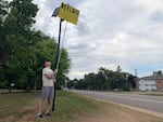 A man uses a long metal pole with a bright yellow sign taped to the end to block the view of a camera mounted on a pole next to a road. The sign says "Deflock Crystal dot com."
