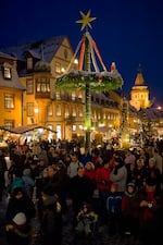 More than 100,000 people typically visit Gegenbach, Germany, (pop. 11,000) in December to view the "world's largest" advent calendar and surrounding Christmas market. This photo shows a crowd of spectarors wearing winter clothes in the town square. It is evening. There are neo-classical buildings behind them and two kiosks selling gifts for Christmas. 