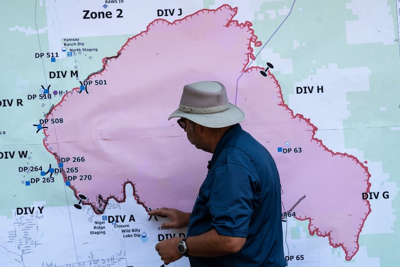 A person stands in front of a wall-sized maps that outlines the boundaries of a wildfire in pink and red.