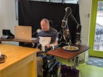 Jonathan Ko is a Seattle attorney with a spinal cord injury. He is also collaborating with robotics researchers at the University of Washington develop a robotic arm that can assist people with motor impairments feed themselves. He brought the Assistive Dexterous Arm into his home in Seattle in Sept. 2024 for a week to feed him meals such as a breakfast of avocado toast, which he is shown doing in this photo, while controlling the device with a mouth-operated joystick.