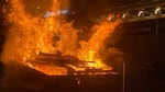 Flames rise out of a Dumpster while the glowing sign of a Eddie Bauer store's sign can be seen in the background behind the fire.