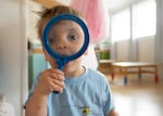 A 2-year-old boy looks through a magnifying glass while facing the camera.