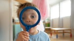 A 2-year-old boy looks through a magnifying glass while facing the camera.