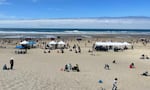 Dozens of people sitting or walking in the sand, with event booths in the foreground, competition plots behind them, and the shore and ocean waves in the background, under a sunny blue sky.