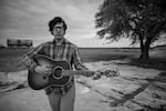 A black and white image of a man standing in a western U.S. landscape with a tree and a blurry structure in the background.