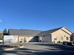 A building that looks like a beige single family home is pictured on a sunny day with no clouds in the sky. A sign next to the driveway into a parking lot says "Phoenix Place."