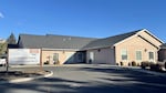 A building that looks like a beige single family home is pictured on a sunny day with no clouds in the sky. A sign next to the driveway into a parking lot says "Phoenix Place."