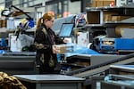 In this photo, a woman at a workstation packs two small cardboard boxes into a large transparent bag.