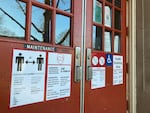 The exterior doors to a school building are plastered in signs. One shows two drawings of people, with a line between them showing they are 6 feet apart. Another shows a person wearing a mask. Others are difficult to read.