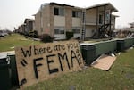 A sheet of plywood with the words "Where are you FEMA?" spray-painted on it leans against a wall in front of an apartment complex with hurricane debris all around.
