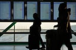 Travelers walk by in profile with screens behind them in an airport terminal.